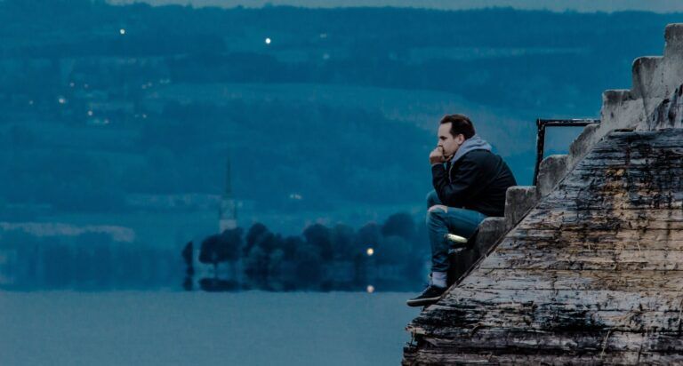 Man sitting at the dock in twilight, thinking about the meaning of life. Lake, headland with church and hills in the background.