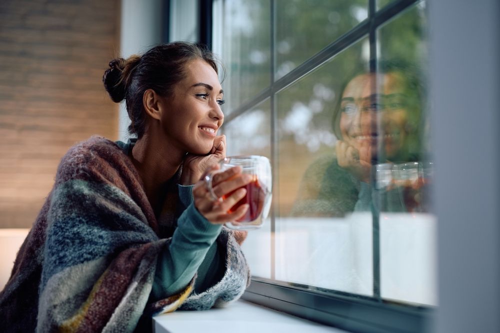 Young smiling woman drinking tea and looking through the window at home