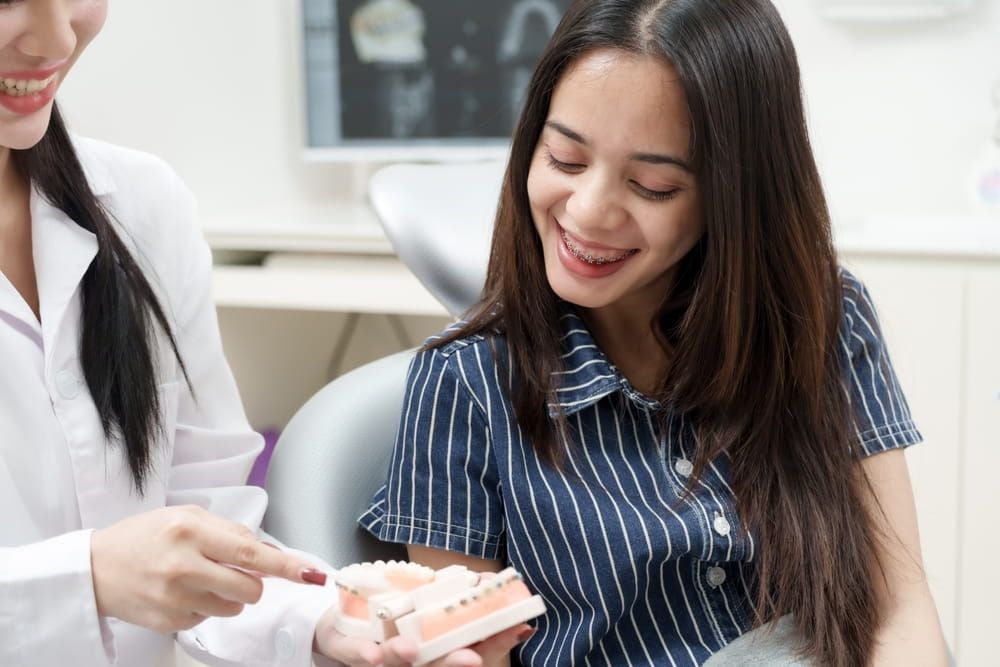 Young adult asian woman with braces sitting in dental chair smiling while listening to asian female dentist showing dental model during orthodontic education in dental clinic