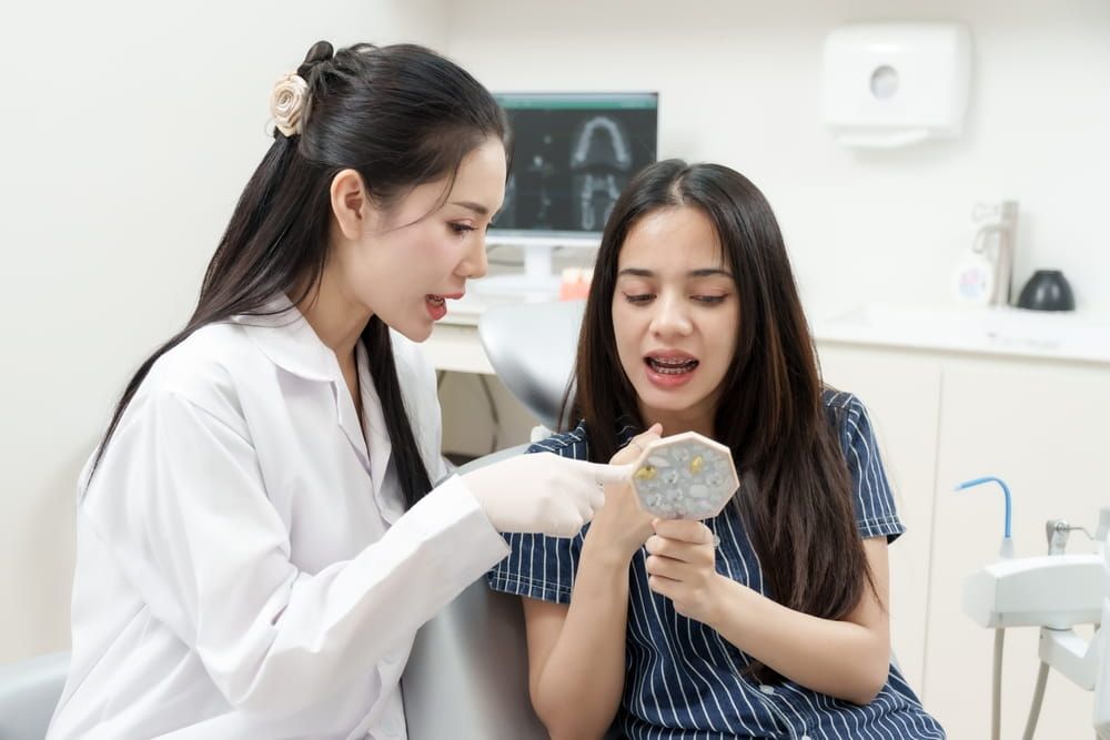 Asian female dentist pointing at tooth model while multiracial female patient with braces listens carefully in dental chair inside modern orthodontic clinic