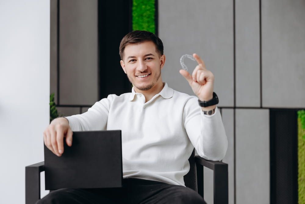 Smiling young man seated in a modern office holding a clear dental aligner, conveying confidence, oral health, and orthodontic treatment in a relaxed, professional environment.