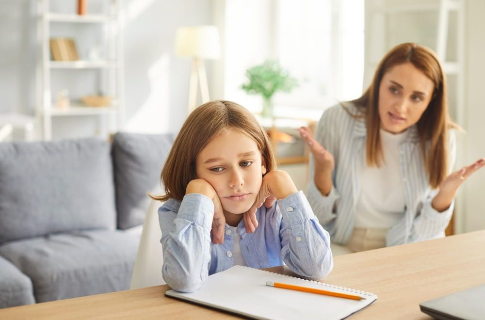 Angry adult mother scolding little child boy for bad school results sitting at desk at home