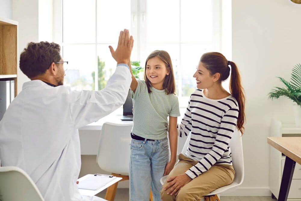 Friendly male pediatrician doctor giving high five to a little child girl patient standing at the doctor's office with her mother during medical examination in clinic.