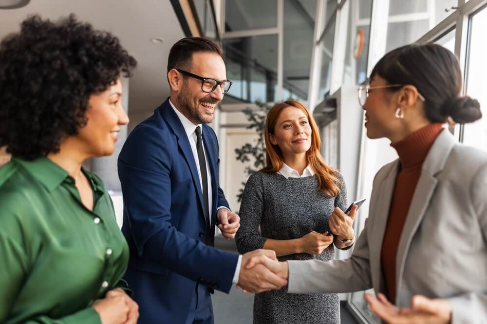 A group of business professionals in formal attire greet each other warmly with a handshake, exuding collaboration and trust, inside a modern and light-filled office environment.