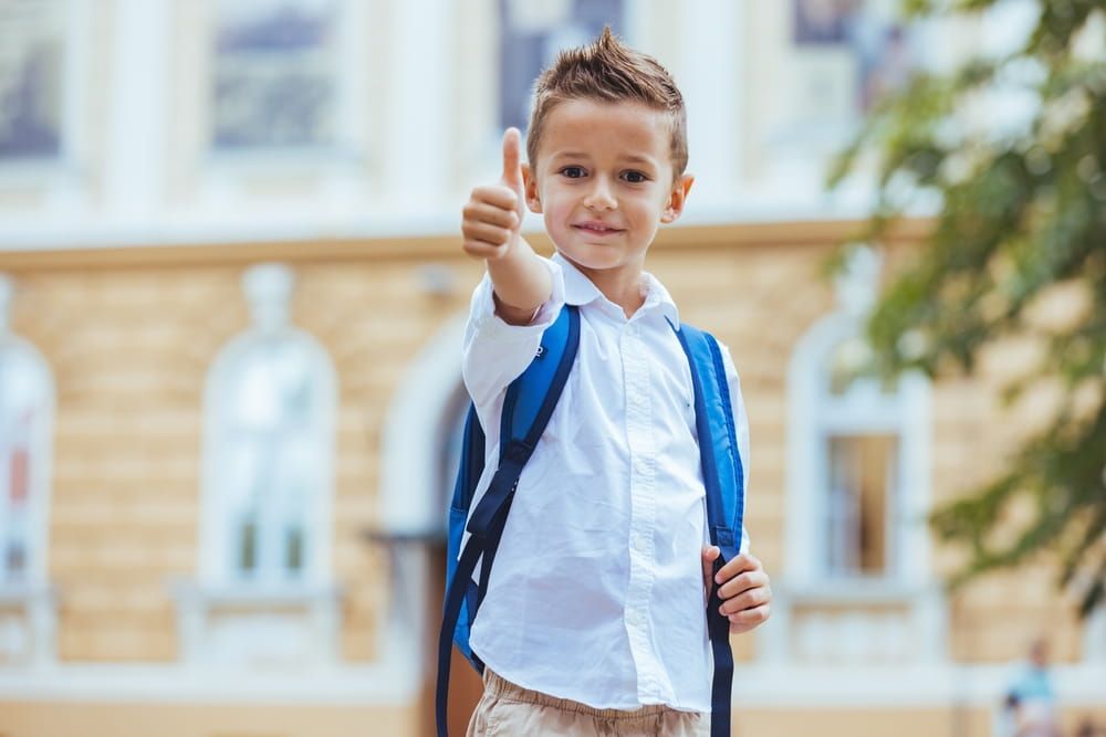 A joyful young Caucasian boy, dressed in a white shirt with a blue backpack, gives a thumbs up, signaling his eagerness for his school day ahead.