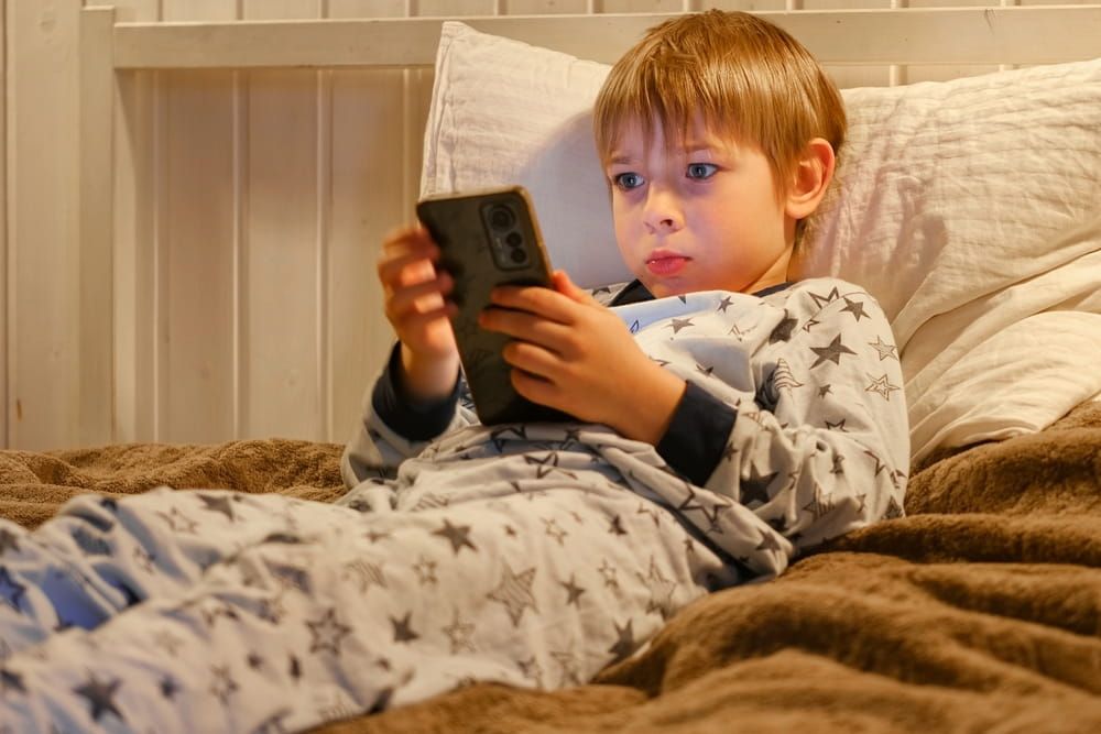 Cute kid boy in pajamas lying in bed watching smartphone, smiling. Children's screen time.
