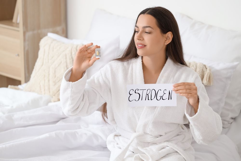 Young woman holding paper with word ESTROGEN and pill in bedroom