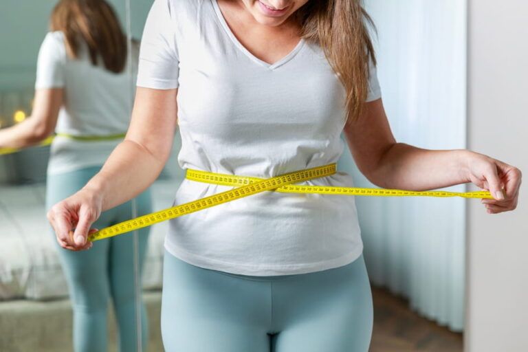 Young woman measuring her waist with tape measure in front of the mirror in her bedroom. Satisfied woman checking her body parameters