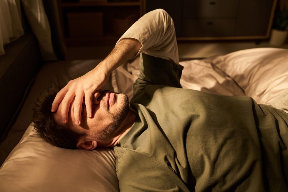 Young man lying on bed covering face with hand appearing stressed or exhausted, resting in bedroom