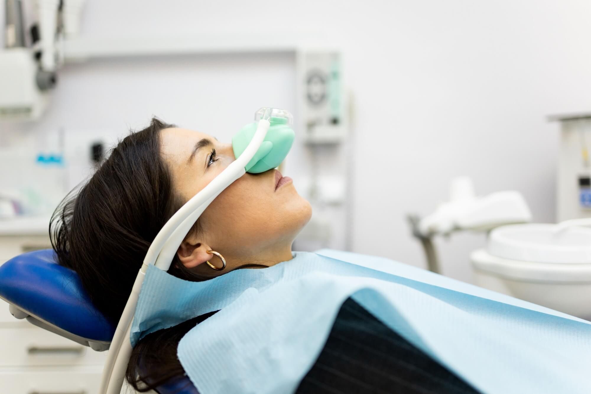 An adult woman sits in a dentist's office wearing a nasal mask to inhale nitrous oxide.