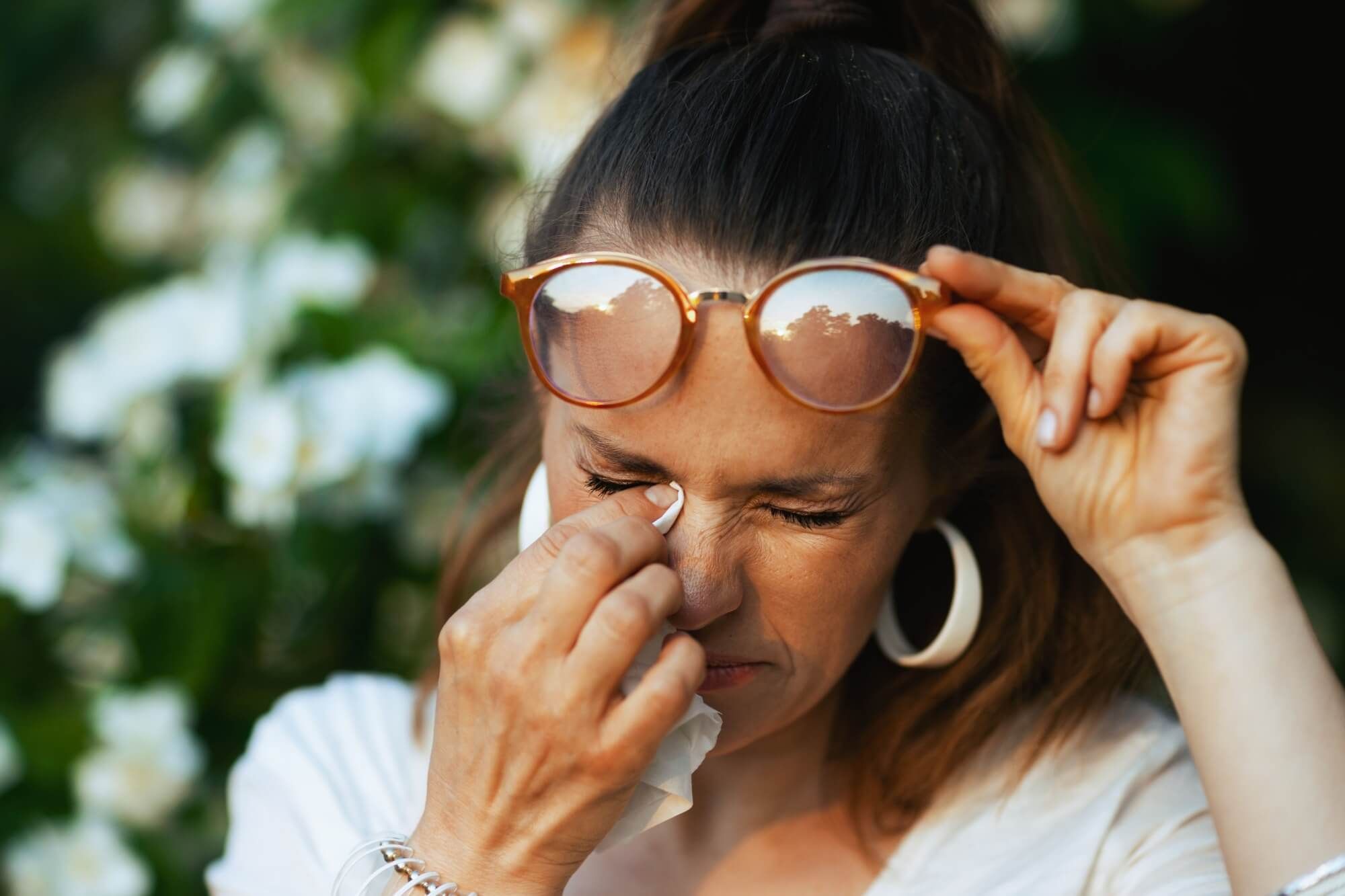 40 years old woman in white shirt with handkerchief and eyeglasses has an allergy attack near flowering tree