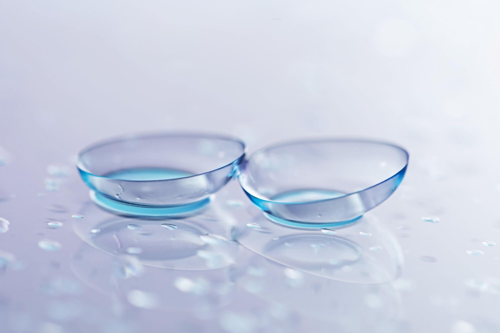 Pair of contact lenses and water drops on light mirror surface, closeup