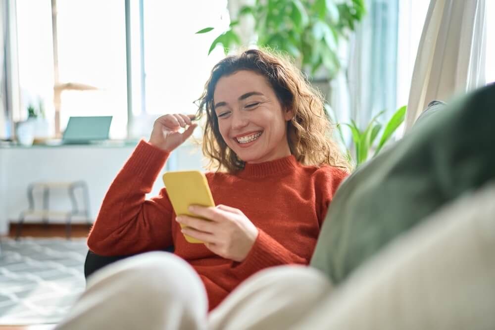 Happy relaxed young woman sitting on couch using cell phone, smiling