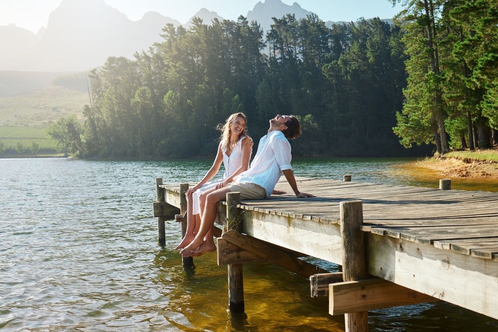 Young couple, lake and jetty with laughing, happiness and bonding with love