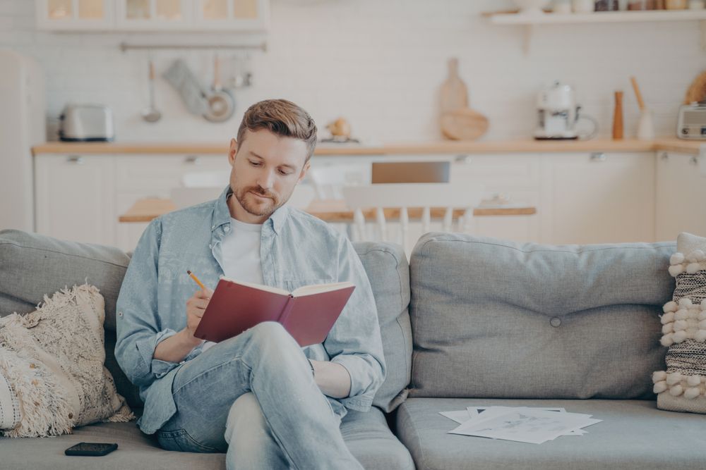 Young man in casual clothes sitting on couch with legs crossed, writing down notes