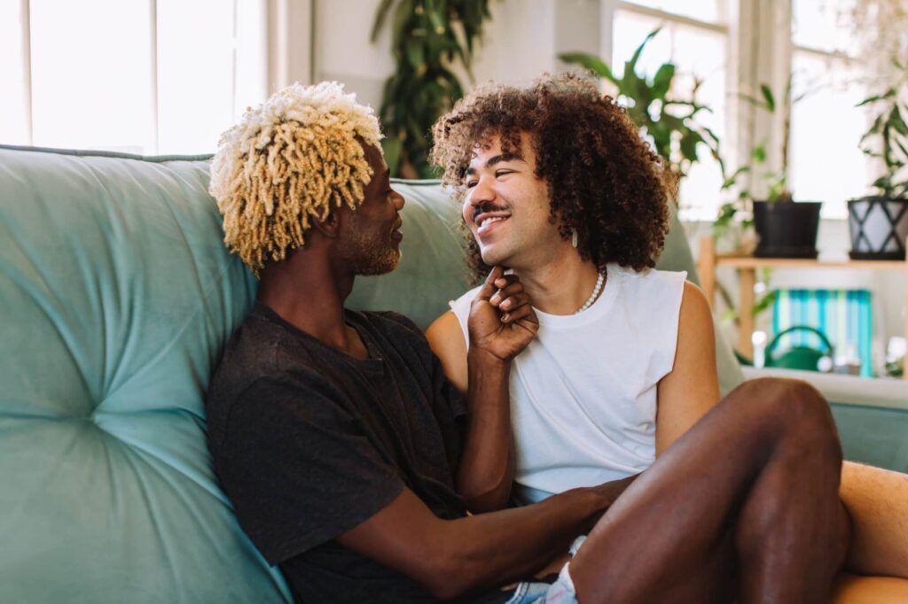 Two affectionate male lovers embracing and smiling at each other while sitting together in their living room