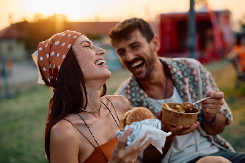 Happy couple of festival goers laughing while enjoying in food at sunset.