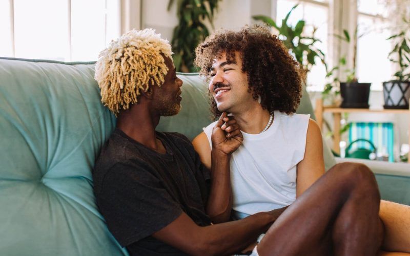 Two affectionate male lovers embracing and smiling at each other while sitting together in their living room
