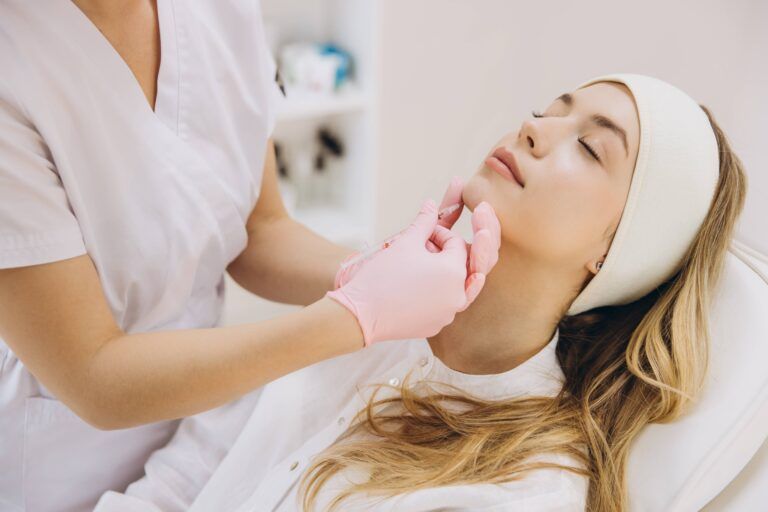 Skilled beautician performing chin augmentation on female patient, lying relaxed during cosmetic facial enhancement procedure in professional clinic setting