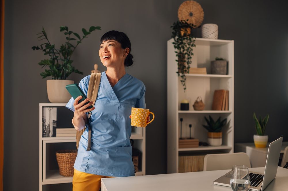 A happy asian nurse wearing blue scrubs is holding a phone and a yellow coffee cup