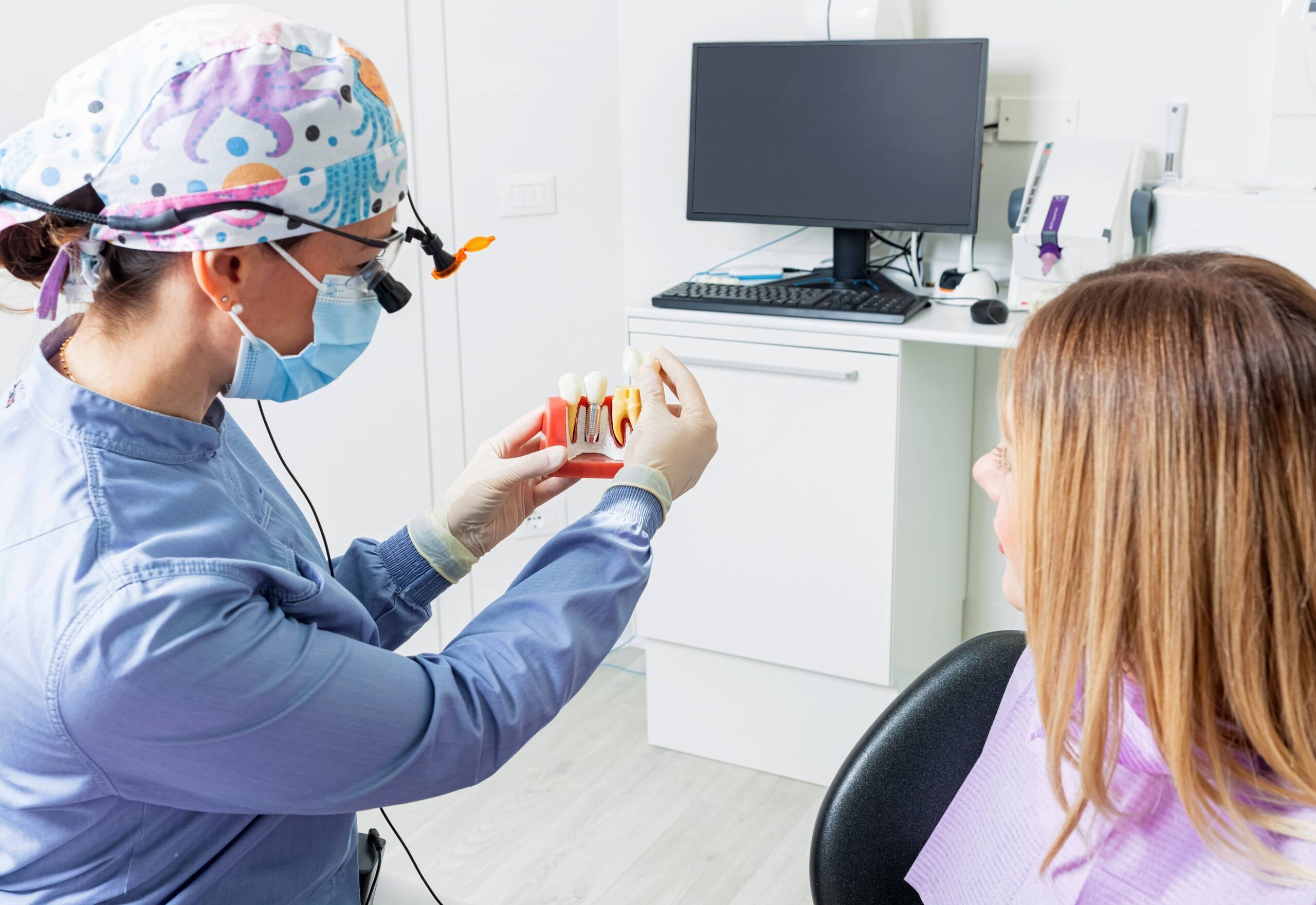 Female dentist explaining dental implant procedure to patient using anatomical model in modern clinic
