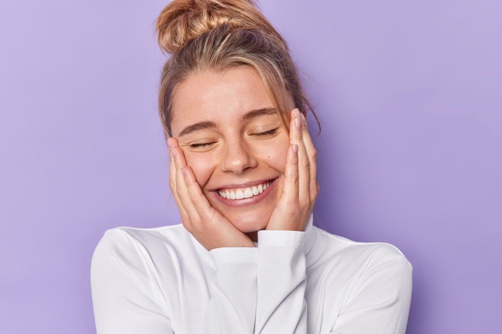 Portrait of cheerful woman keeps eyes closed smiles broadly has combed hair wears white casual jumper isolated over purple studio background.