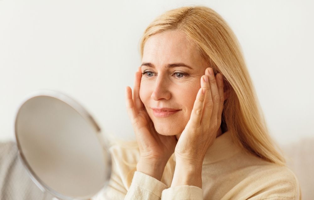 A blonde woman with a gentle smile checks her skin in the mirror, holding her hands to her cheeks.