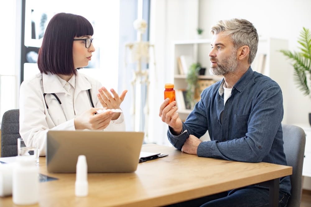 Female general practitioner in white uniform communicating with mature patient holding bottle of medication in new clinic