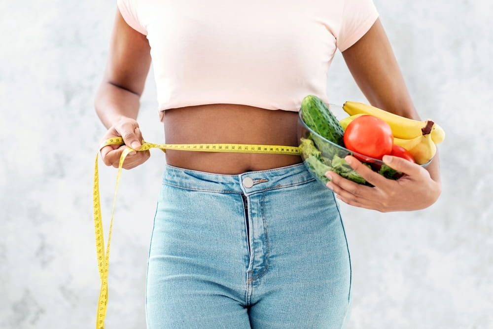 Diet for weight loss. Unrecognizable black woman holding bowl of fruits and veggies with tape measure