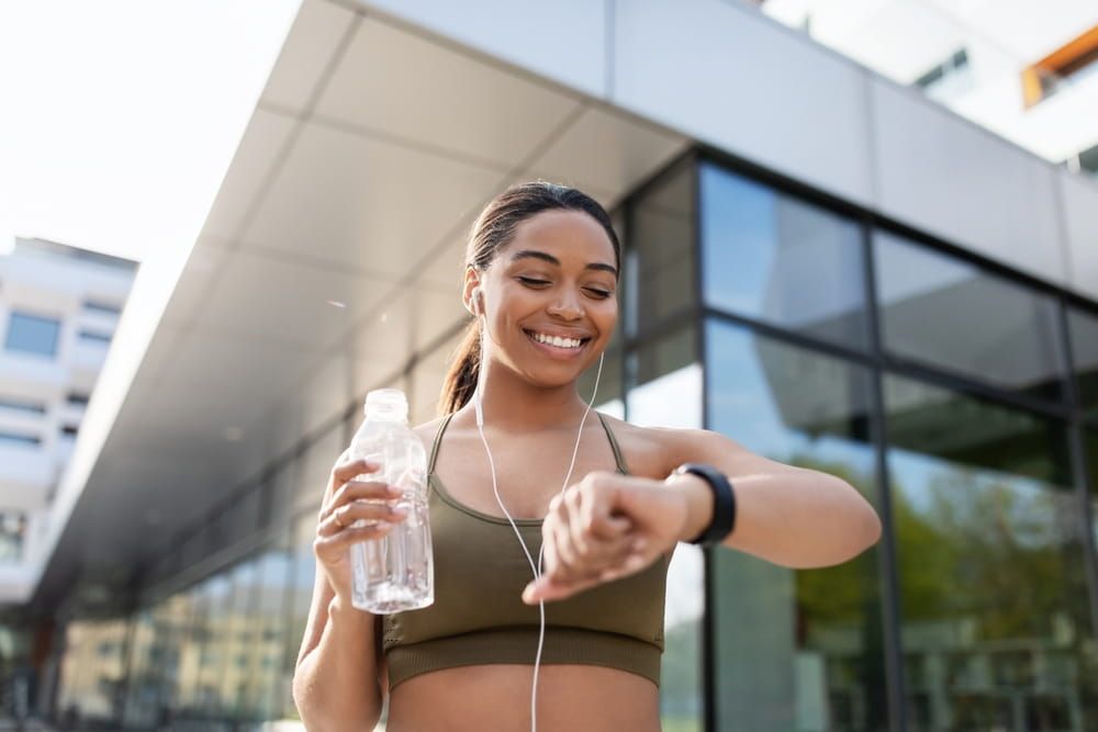 Young black woman looking at smartwatch, tracking fitness activity, drinking water on training outdoor