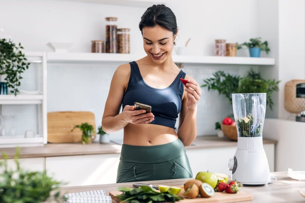 athletic woman using mobile phone while preparing smoothie in the kitche