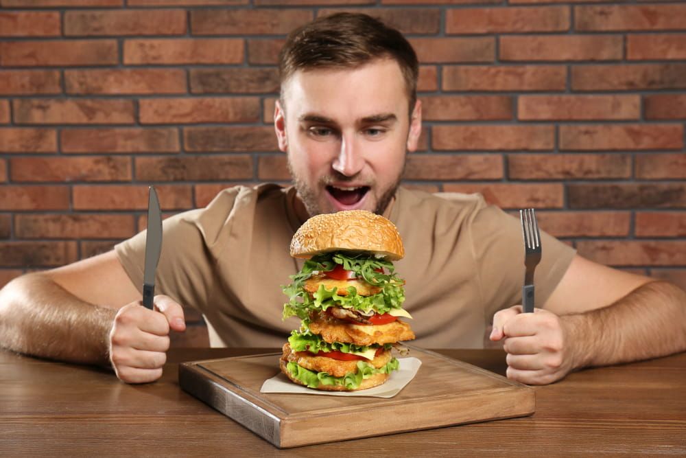 Young hungry man with cutlery eating huge burger at table