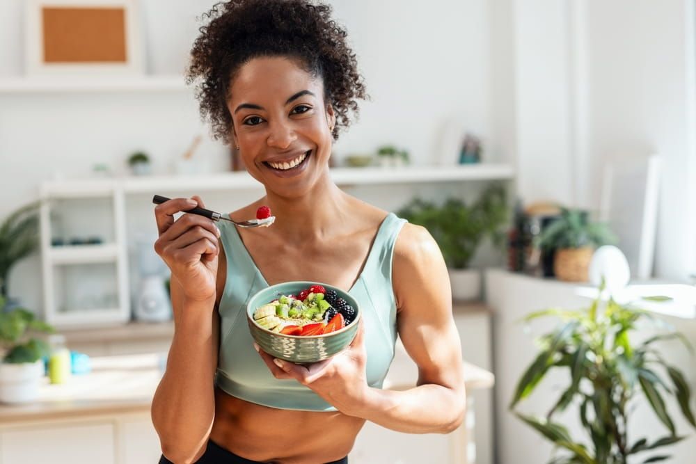 athletic woman eating a healthy fruit bowl while looking at camera in the kitchen at home