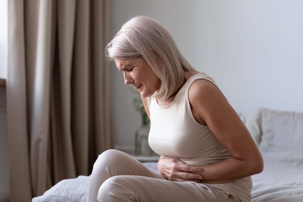 Middle-aged woman sitting on bed feels unhealthy touch stomach suffers from severe crampy abdominal pain