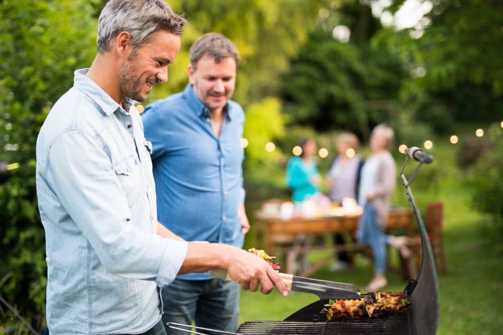 in a summer evening, two men in their forties prepares a barbecue for friends gathered around a table in the garden