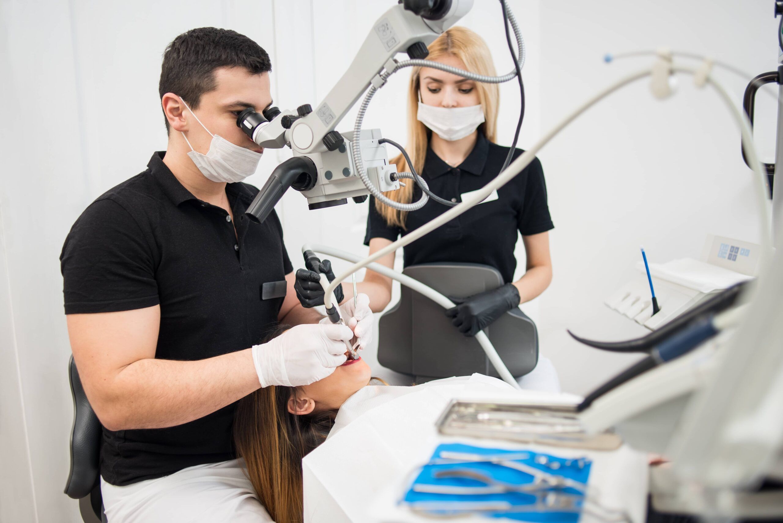 Male dentist and female assistant treating patient teeth with dental tools - microscope, mirror and drill at dental clinic office
