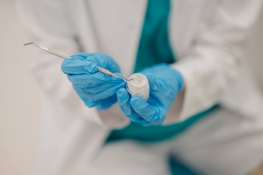 Dentist wearing blue gloves picking up dental filling material from a small container using a dental tool