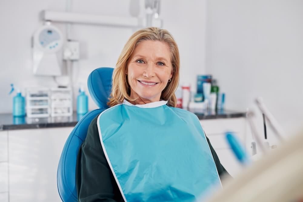 Old beautiful lady smiling while seated on dental chair wearing a blue bib for dental implant