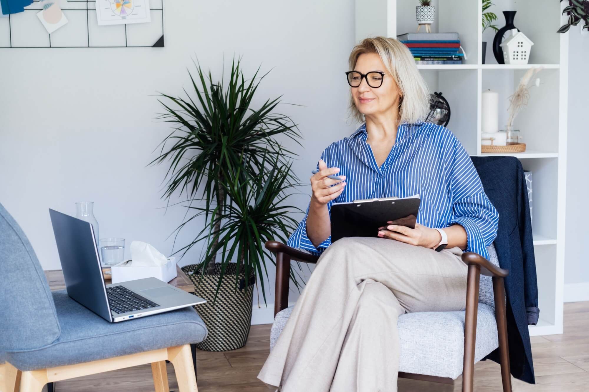 Middle aged female Psychologist having video call consultation on laptop while sitting on armchair In office.