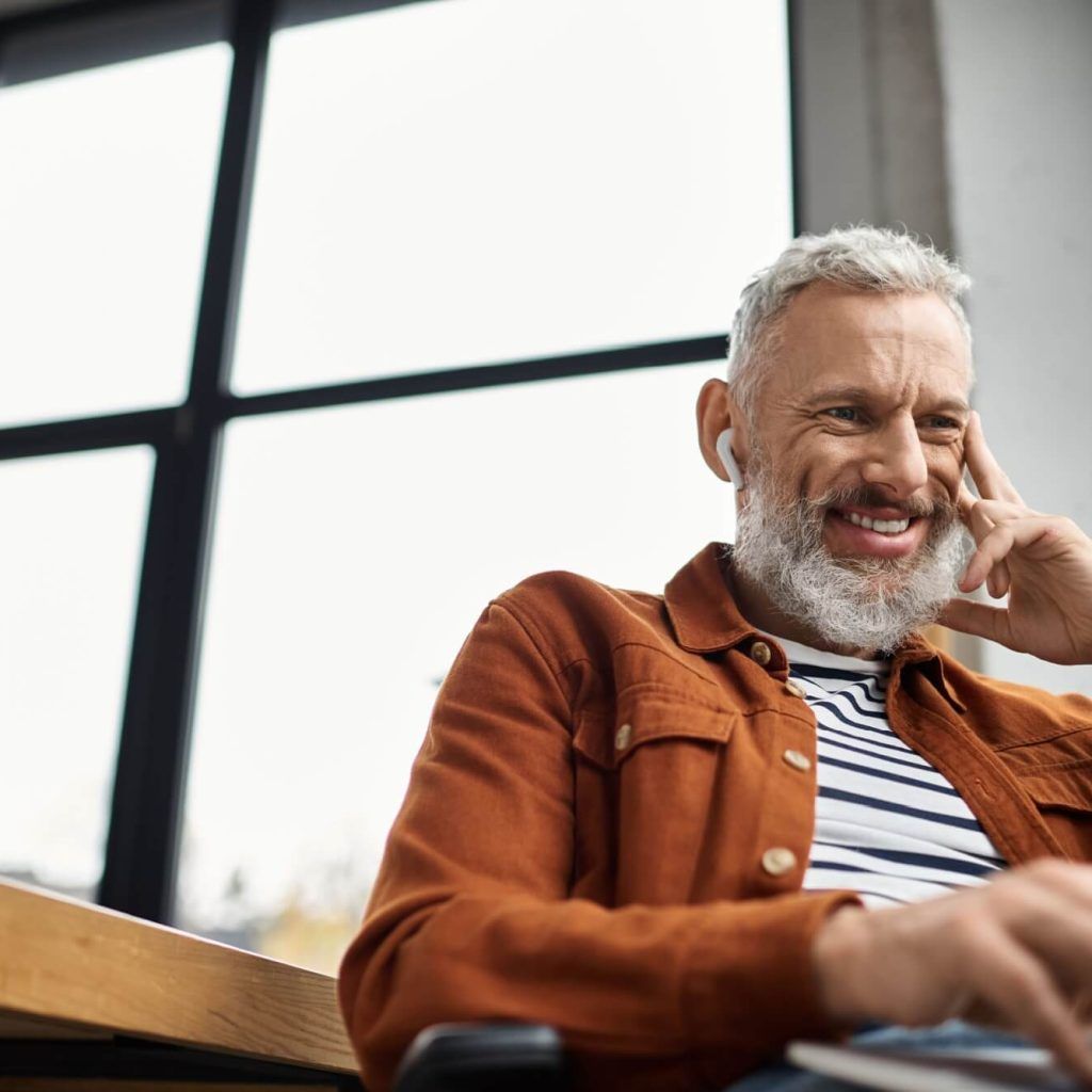A man with a beard working happily at his laptop in a sunlit office, wearing wireless earbuds.