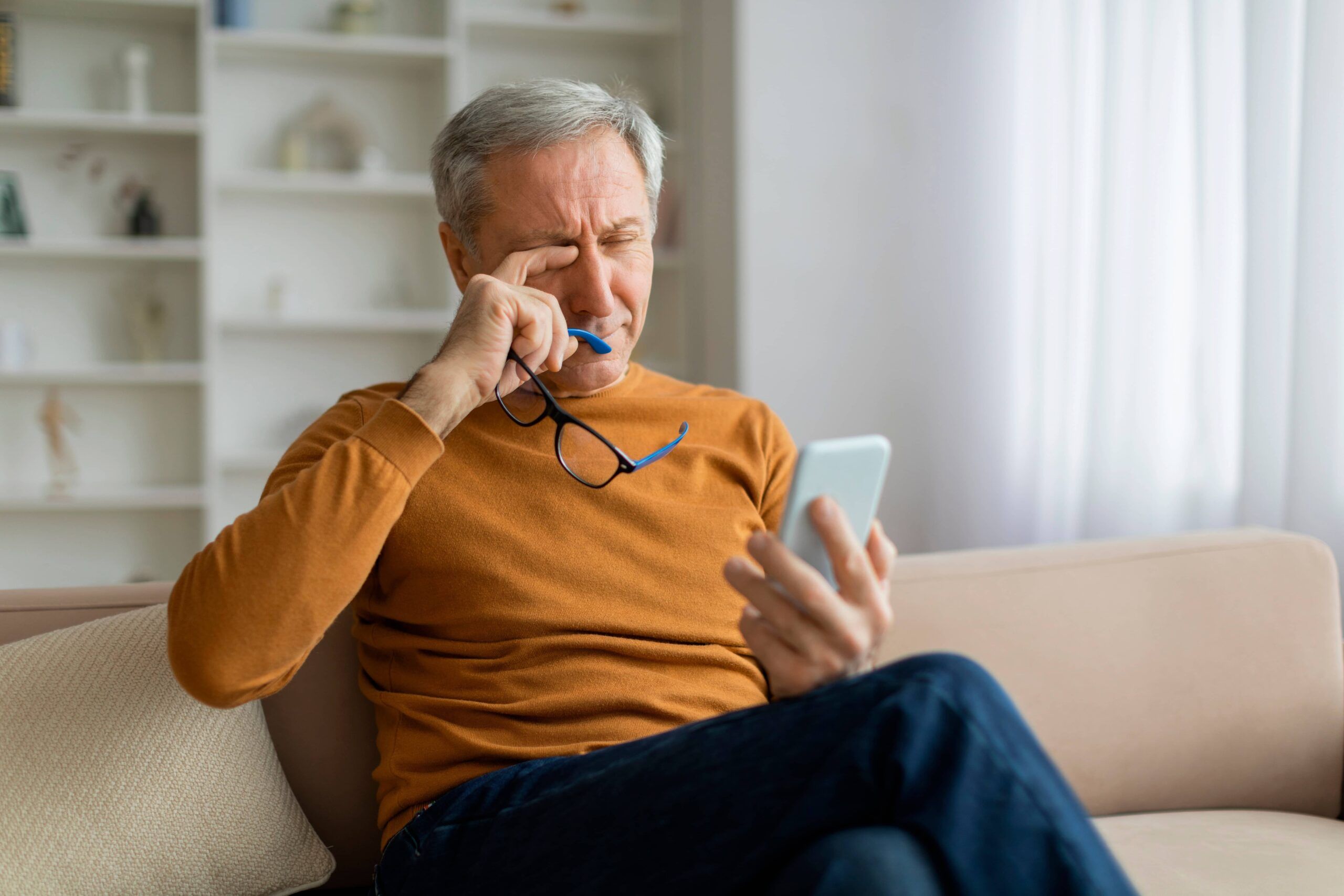 senior man sitting on couch in cozy living room, holding eyeglasses, rubbing his eyes