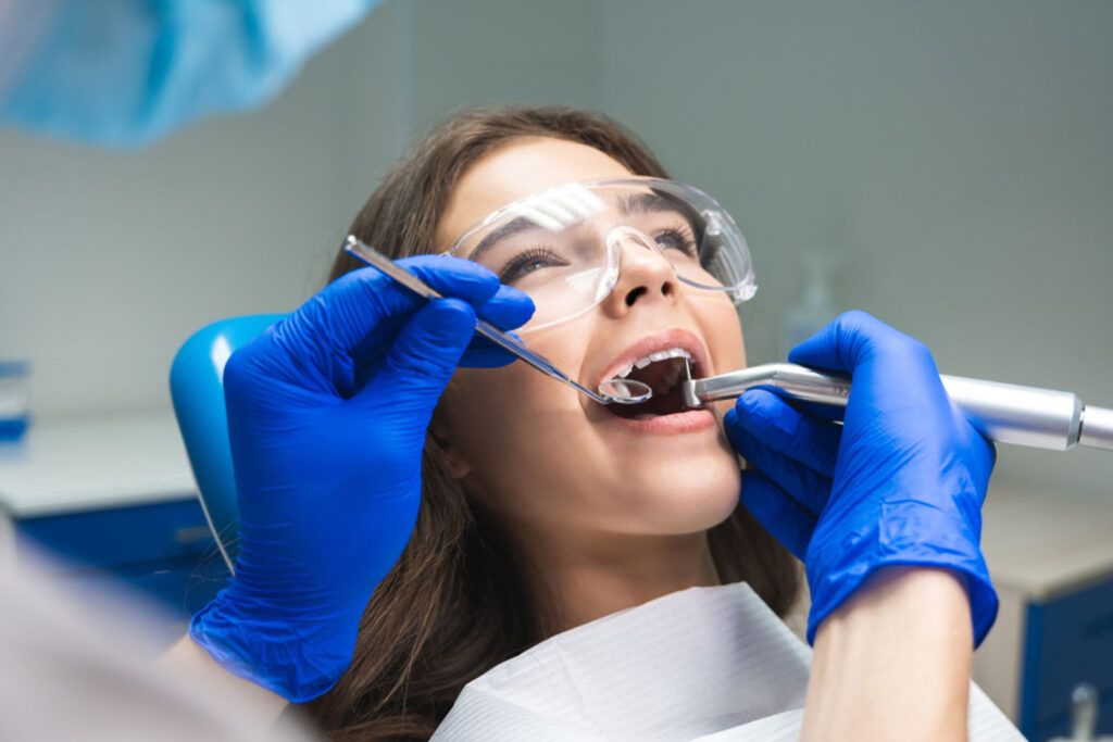 dentist in mask filling the patient's root canal while she is lying on dental chair wearing safety glasses under the medical lamp in dental office