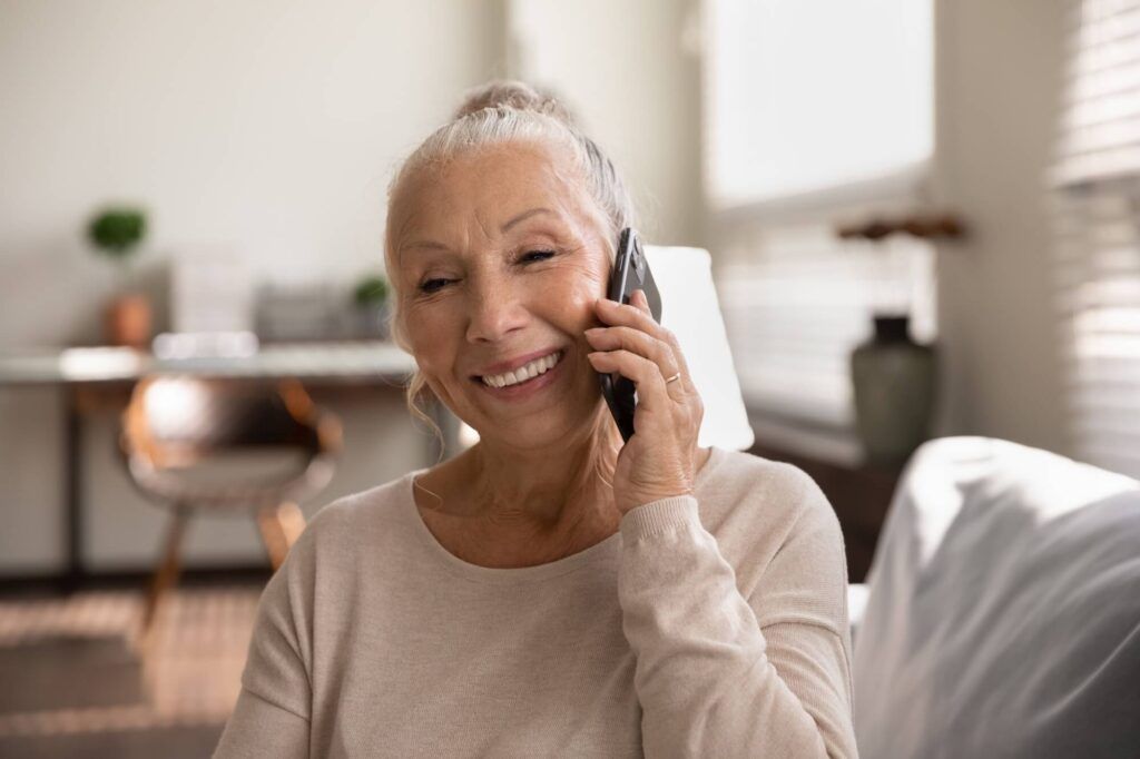 Happy elderly lady talking on cellphone, making call from home, speaking to family on mobile phone with toothy smile