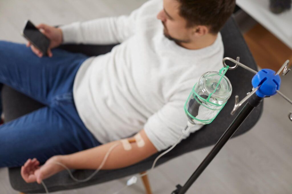 Top view photo of a man sitting in the chair and using the smartphone while receiving IV drip infusion and vitamin therapy