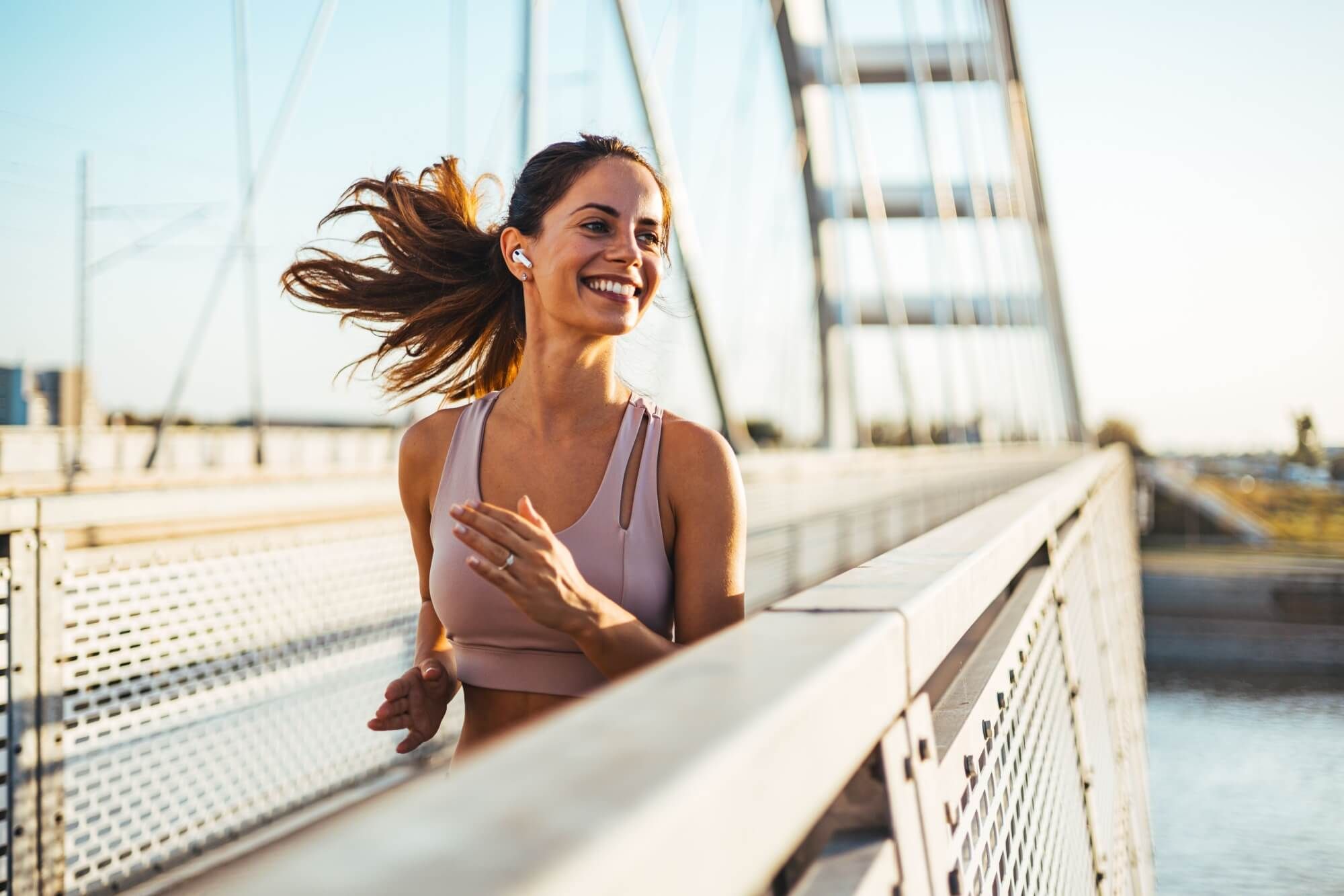 Athletic woman jogging with a bright smile on a bridge under a clear sky. Her hair flows in the wind, emphasizing her joyful and energetic attitude towards fitness and outdoor exercise.
