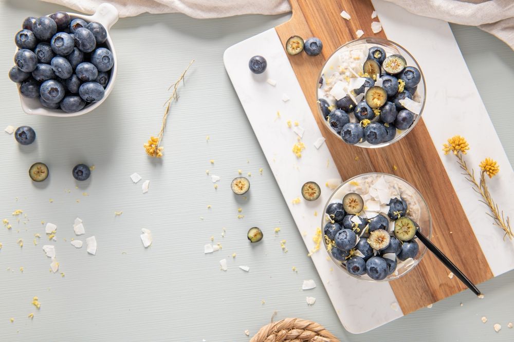 Overnight Oats topped with blueberries, lemon zest and coconut shavings on a light blue wooden counter
