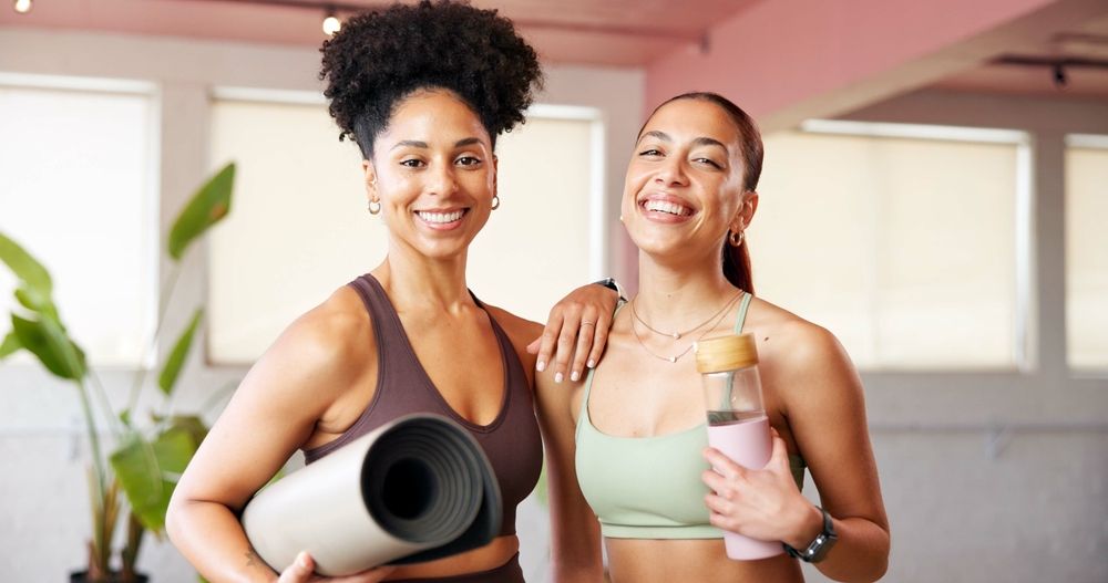 two happy women smiling and holding a water bottle and yoga mat