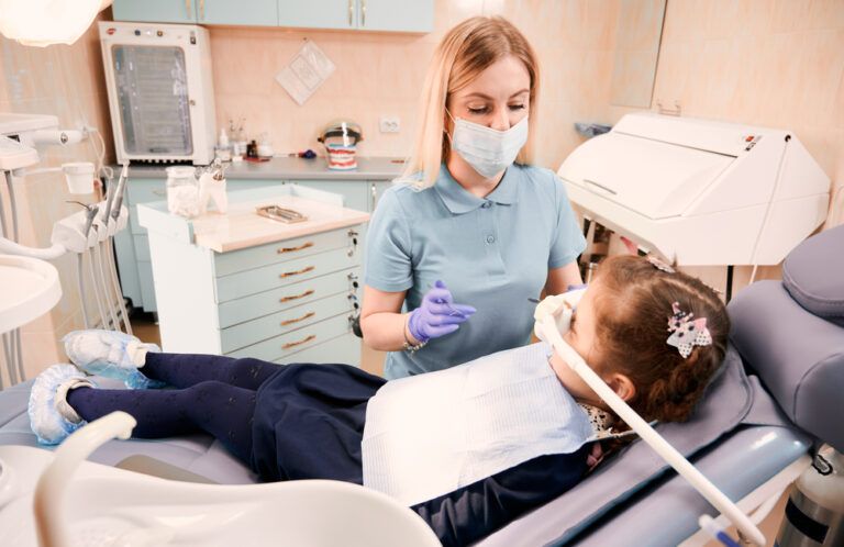 Young woman dentist in medical face mask holding dental instruments while little girl lying in dental chair