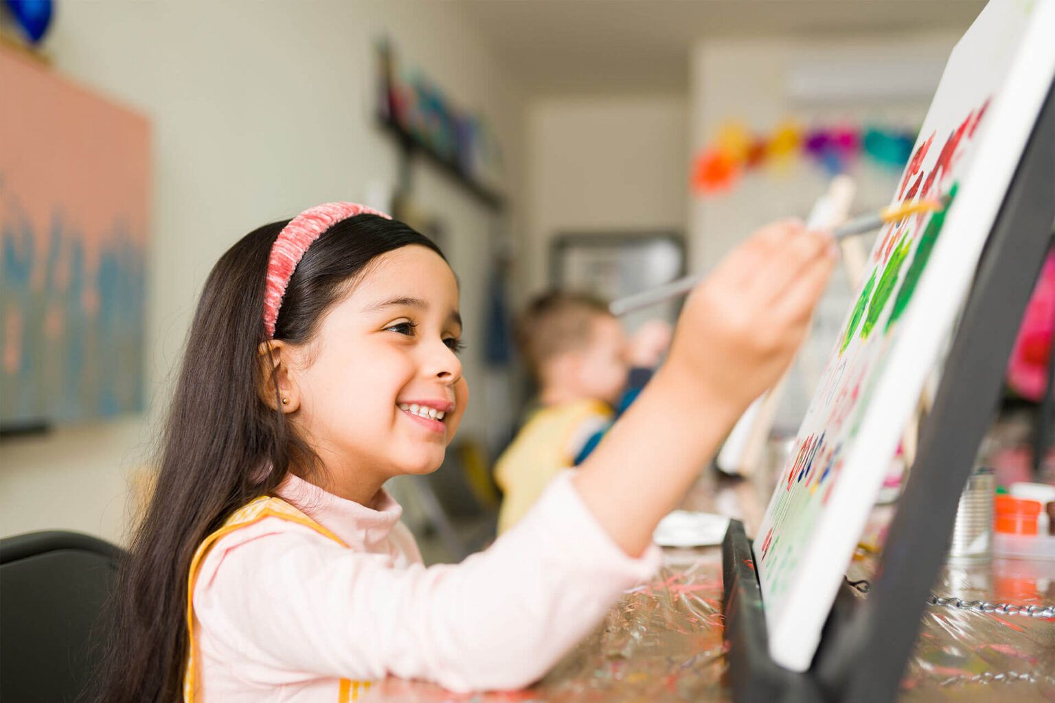 Side view of a happy latin girl kid painting a colorful landscape with a brush on a canvas