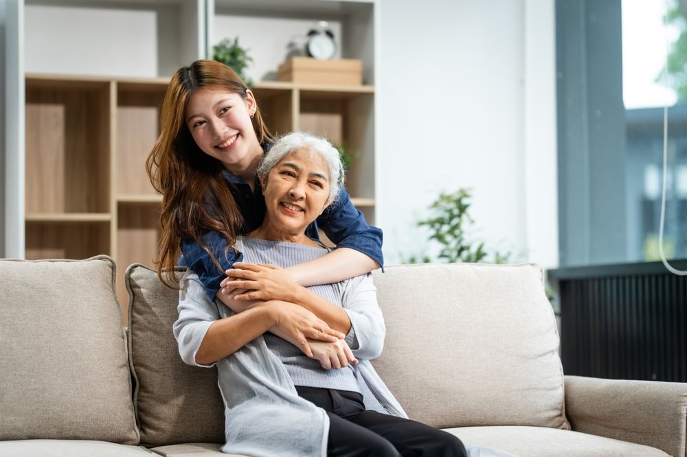 A mature mom and a young Asian woman, mother and daughter, sit together on a sofa in their living room